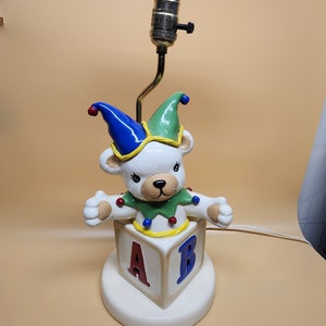 May include: A ceramic table lamp shaped like a teddy bear wearing a jester hat and sitting on top of a block with the letters A and B. The lamp is white with green, blue, red, and yellow accents.