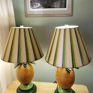 Puede incluir: Un par de lámparas de mesa con forma de piña con pantallas de lámparas a rayas verdes y beige. Las lámparas están sobre una superficie de madera.