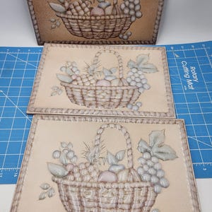 May include: Three decorative ceramic tiles with a raised design of a basket filled with fruit and grapes. The tiles are beige with a rope-like border.