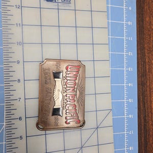 以下が含まれることがあります： 赤と白で「UNION PACIFIC RAILROAD COMPANY」の文字が入った長方形の金属製ベルトバックル。 バックルには握手のグラフィックと1860年と1885年の年号が描かれています。 バックルの長さは約7.6cmです。