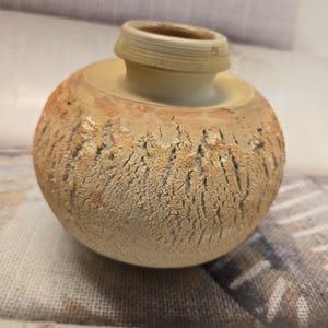 Puede incluir: Un pequeño jarrón de cerámica redondo con una superficie texturizada y agrietada en tonos beige y bronceado. El jarrón presenta un cuello liso y elevado con un borde ancho y plano. El cuerpo tiene un aspecto desgastado con grietas verticales.