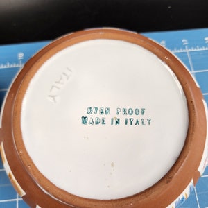 May include: The bottom of a white ceramic dish with a brown rim. The dish has a colorful pattern and the text "OVEN PROOF MADE IN ITALY" is printed on the bottom.