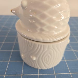May include: White ceramic hedgehog figurine with a gold accent on its nose. The hedgehog is sitting on top of a white ceramic tree stump with a textured wood grain design.