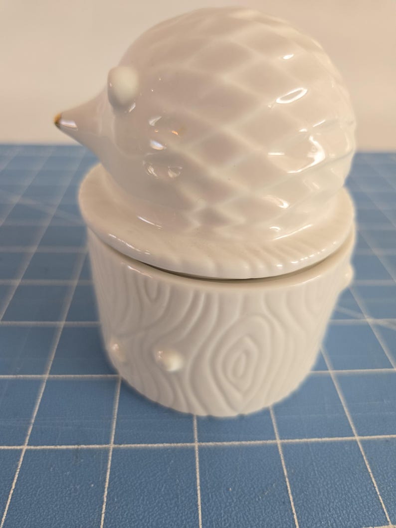 May include: White ceramic hedgehog figurine with a gold accent on its nose. The hedgehog is sitting on top of a white ceramic tree stump with a textured wood grain design.