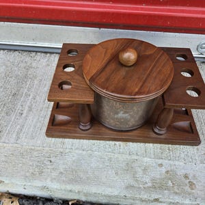 May include: A wooden tobacco pipe stand and humidor. The stand is dark brown with cutouts for pipes and a central container. The humidor has a wooden lid with a small knob. The container is clear with a silver-colored interior.
