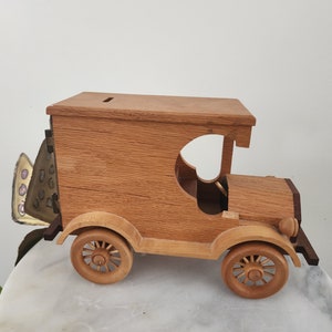 May include: Wooden toy car shaped like a vintage delivery truck with a slot in the roof for coins. The car is painted in a natural wood finish and has a detailed design.