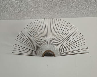 May include: Metal wall art depicting a stylized sunburst with radiating lines. Three birds are silhouetted against the sun. The artwork is mounted on a white wall, creating a striking contrast.