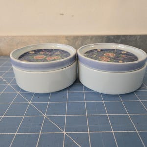 Puede incluir: Dos cajas redondas de cerámica para guardar pequeños objetos con diseños florales. Las cajas son blancas con un borde azul claro y un patrón floral en la tapa. El patrón floral es en tonos de rosa, azul y verde.