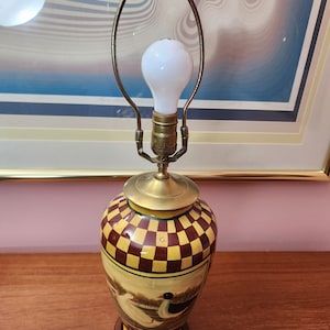 May include: A ceramic table lamp with a gold-colored base and a checkered design. The lampshade is made of metal and has a simple, curved design. The lamp is lit with a white light bulb.
