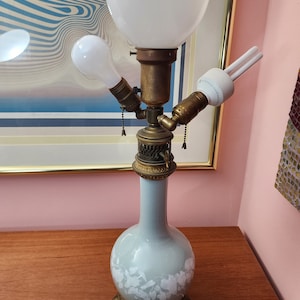 May include: A three-light table lamp with a white ceramic base decorated with a floral pattern. The lamp has a white glass shade and three brass light fixtures.