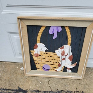 May include: A framed wooden wall art piece featuring two dogs in a basket. The dogs are white with brown spots and are painted in a cartoon style. The basket is woven and has a purple bow on top. The background is black with a wood grain texture. The art is signed "LeGroot".