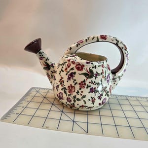 Może przedstawiać: Mała, ceramiczna konewka z kwiatowym wzorem w odcieniach czerwieni, fioletu i zieleni na kremowym tle. Dzióbek i uchwyt są ciemnobrązowe. Konewka ma około 15 cm długości.