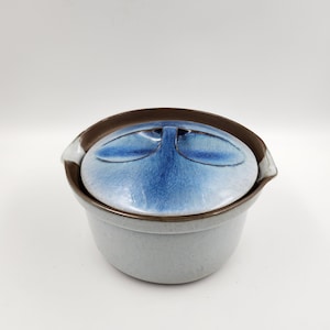 May include: A ceramic casserole dish with a lid. The dish is light gray with a brown rim and handles. The lid is blue with a darker blue center and a brown handle. The dish is on a white background.