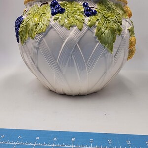 Op de afbeelding: Witte keramische plantenbak met een geweven ontwerp en wijnstokaccenten. De plantenbak heeft een gele rand en is versierd met groene bladeren en blauwe druiven.
