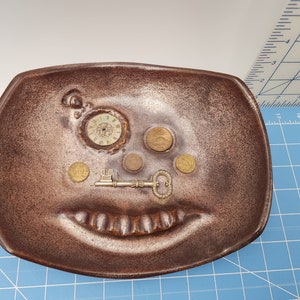 May include: A brown ceramic dish with a face design. It features a clock, coins, and a key. The dish has a textured surface and a whimsical, artistic aesthetic. A unique decorative item.