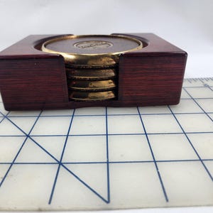 May include: A set of coasters with a dark wood holder. The coasters are gold-toned with a brown center. The holder is a dark reddish-brown color. The coasters are stacked inside the holder.