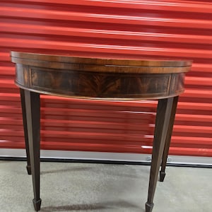 May include: A vintage, half-round wooden table with a dark brown finish. The table features a curved apron with decorative wood grain and slender, tapered legs. The tabletop is a lighter shade of brown.