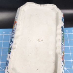 May include: White ceramic planter with a floral design in blue, orange, yellow, and green. The planter has a rectangular shape with a slightly raised rim.