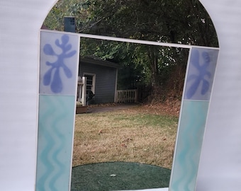 May include: A white arched mirror with a decorative frame. The frame is made of glass tiles with blue and green abstract designs. The mirror reflects a green lawn and a gray shed.