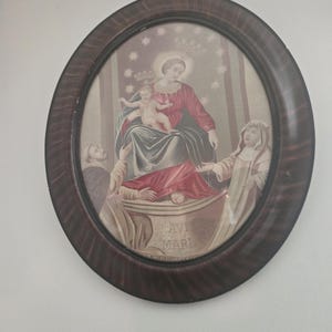 Può includere: Una stampa ovale incorniciata raffigurante una scena religiosa. L'immagine presenta una donna in una veste rossa e blu che tiene in braccio un bambino, con due figure sottostanti. La cornice è marrone scuro con un motivo a venature del legno. Le parole "Ave Maria" sono visibili.