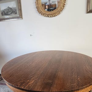Könnte beinhalten: Ein runder Holztisch mit einer dunkelbraunen Oberfläche. Der Tisch hat einen Sockel mit einem geschnitzten Design. Der Tisch steht in einem Raum mit einem goldenen Spiegel an der Wand.