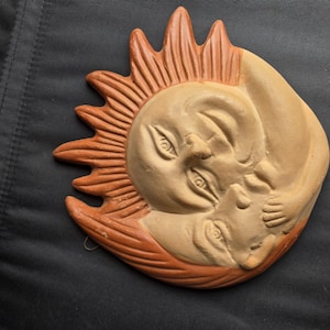 May include: A terracotta wall plaque featuring a relief sculpture of a sun and moon face intertwined. The sun has radiating rays and a light tan face, while the moon is depicted in a darker clay tone with a closed eye and hand resting near the sun.