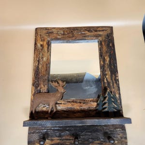 May include: A rustic wooden mirror with a shelf and hooks. The mirror has a brown wooden frame with a distressed finish. The shelf is made of dark wood and has two hooks on the bottom. There is a wooden deer and a pine tree on the shelf.