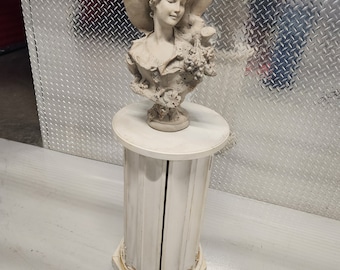 May include: A white pedestal with a white painted finish and a decorative, sculpted bust of a woman wearing a hat. The bust is made of plaster or ceramic and has a detailed design.