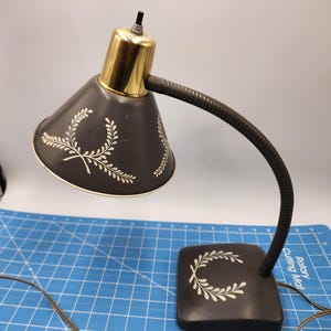 May include: A vintage black desk lamp with a gold-colored top and a flexible gooseneck. The lampshade has a white floral design and the base has a white wreath design.