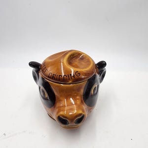 May include: A brown ceramic cow head shaped butter dish with black eyes and a black nose. The lid has the words "Cheese Dripping" written on it.