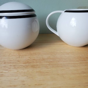Peut inclure: Un ensemble de crémier et de sucrier en céramique blanche avec une bordure noire. Le pichet à crème a une poignée et le sucrier a un couvercle.