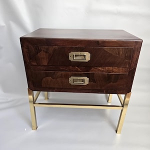 Op de afbeelding: Een donkerbruin houten nachtkastje met twee lades en messing hardware. Het nachtkastje wordt ondersteund door een goudkleurig metalen frame. De houtnerf is zichtbaar op de lades.