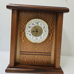 May include: A wooden mantel clock with a light-colored face and gold-colored hands. The clock face has black numerals and a gold-colored center. The clock is housed in a dark brown wooden frame with a lighter wood panel in the center.
