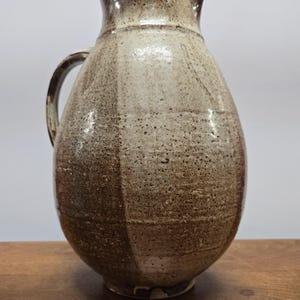 May include: A brown and white ceramic jug with a rounded body and a handle. The jug has a speckled glaze and a slightly uneven surface.