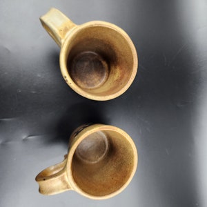 May include: Two handmade ceramic mugs with a rustic, earthy aesthetic. The mugs are a warm, beige color with a textured surface and a dark brown interior. Each mug has a curved handle for easy gripping.