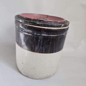 May include: A vintage ceramic crock with a lid. The crock is cylindrical, with a white base and a dark brown upper section. The lid is pink and fits snugly on top. The piece has a rustic, aged appearance.
