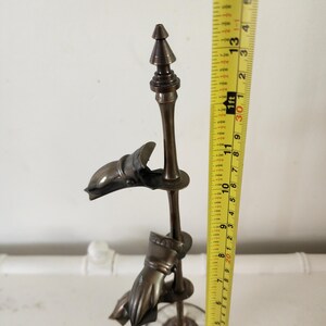 Könnte beinhalten: Eine bronzefarbene Metallskulptur mit zwei Händen, die eine zentrale Stange umfassen. Die Skulptur ist etwa 33 cm hoch, hat einen dekorativen Sockel und eine spitze Spitze. Die Hände sind detailliert.