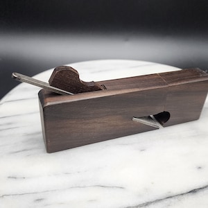 May include: A dark wood hand plane with a metal blade. The plane is on a white marble surface.