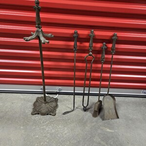 May include: A set of five antique fireplace tools with ornate metal handles. The tools include a poker, tongs, shovel, and brush. The tools are mounted on a decorative stand with a scrolled design.
