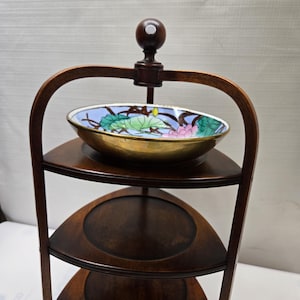 May include: A three-tiered wooden serving stand with a curved design. The top tier has a decorative bowl with a floral pattern in shades of blue, pink, and green. The stand is made of dark wood and has a simple, elegant design.