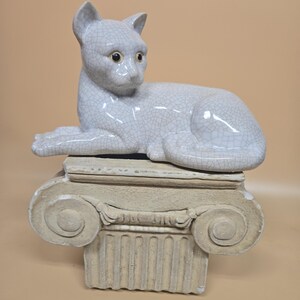 May include: A light gray ceramic cat statue with a crackle glaze finish, posed lying down on a decorative column. The cat has detailed features and is resting on a beige, classical-style column. The statue is a decorative home accent.