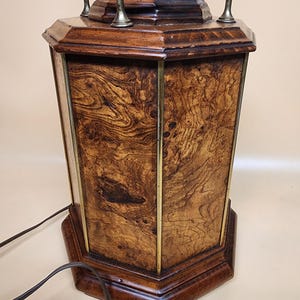 May include: Octagonal wooden base with a brass top and a decorative ring. The base has a wood grain pattern and is dark brown in color. The brass top has a smooth, shiny finish.