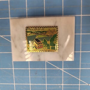 May include: A gold-colored metal souvenir pin with a white marble background. The pin features a depiction of a red and brown trolley car traveling up a hill with a cityscape in the background. The pin has a gold border and the words "Pittsburgh" in gold letters.