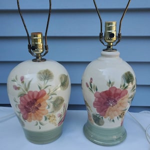May include: A pair of ceramic table lamps with floral designs. The lamps have a white base with green accents and feature pink, orange, and yellow flowers.