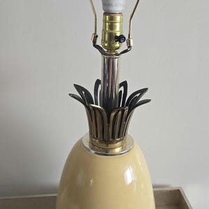 May include: A yellow ceramic pineapple table lamp with a brass base and a black metal pineapple top. The lamp has a white light bulb and a brass switch on the cord.