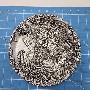 May include: A silver metal plate featuring an embossed design of a jungle scene. The scene includes a jaguar, birds, and foliage.
