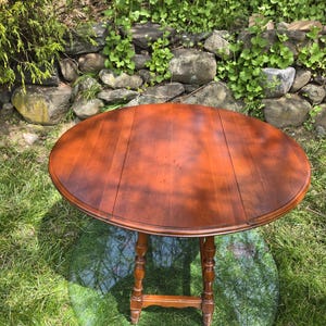 May include: A round, wooden drop-leaf table with a rich, reddish-brown finish. The table has a turned pedestal base and is set on a clear, round glass surface. The table is approximately 36 inches in diameter when fully extended.
