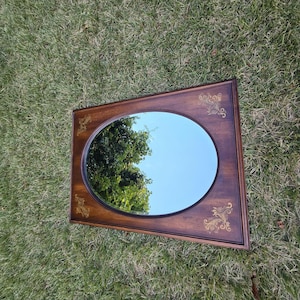 Pode incluir: Um espelho retangular de madeira com uma superfície oval. A moldura castanha escura tem detalhes decorativos dourados em cada canto. O espelho reflete um céu azul e árvores verdes.