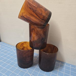 Puede incluir: Cuatro vasos marrones con un patrón de vetas de madera arremolinadas. Los vasos están apilados, con uno inclinado encima de los otros. Los vasos tienen una base marrón oscuro y un interior marrón más claro.