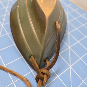 May include: A leaf-shaped ceramic pendant in shades of green and gold, with a textured surface. Brown leather cord is knotted through the pendant's holes, creating a necklace or decorative element.